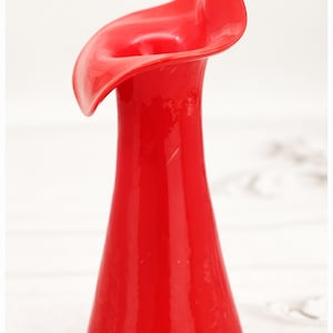 May include: A bright red glass vase with a distinctive, flowing form. The vase features a flared top that curves elegantly, reminiscent of a calla lily. The glossy finish and striking colour make it a decorative piece.