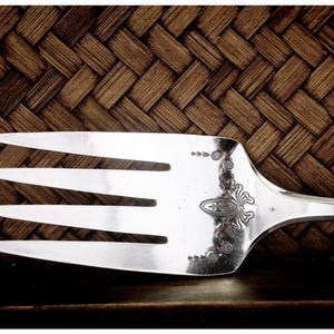 Puede incluir: Un tenedor de servir plateado con cuatro púas, con detalles grabados ornamentales en el mango. El tenedor se encuentra sobre un fondo marrón texturizado y tejido, lo que sugiere una estética rústica o vintage. Las púas son afiladas y pulidas.