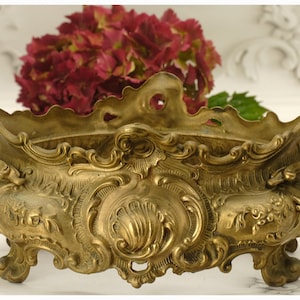 以下が含まれることがあります： 精巧な装飾が施されたアンティーク真鍮製のプランター。花柄と渦巻き模様が特徴です。プランターには2つのハンドルがあり、4つの小さな足の上に置かれています。プランターの中には、深紅色の花の束が見え、生け花に使用されていることがわかります。