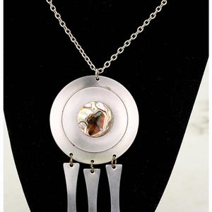 Op de afbeelding: Zilverkleurige hangerketting met een cirkelvormig ontwerp. De hanger heeft een centraal abalone schelp accent omgeven door concentrische cirkels. Drie langwerpige, rechthoekige bedels hangen eronder, bevestigd aan een zilveren ketting.