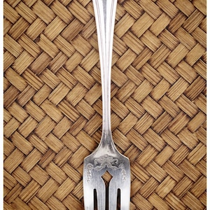 May include: A silver fork with a decorative handle and three tines. The fork has an ornate design on the handle and the tines are pointed. The background is a woven pattern in shades of brown.
