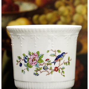 May include: White ceramic planter with a floral and bird design. The planter features a detailed pattern of pink, blue, and red flowers with a blue bird perched on a branch. The rim has a decorative embossed design.