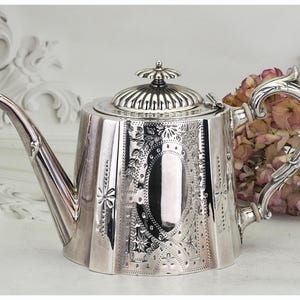 May include: A silver teapot with detailed floral and geometric engravings. The teapot has a curved spout, a decorative handle, and a lid with a small finial. The polished surface reflects light, showcasing the craftsmanship of this antique tea set.