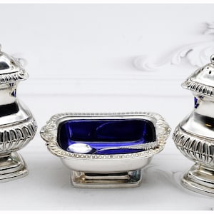 Puede incluir: Un juego de salero y pimentero de plata con un plato de vidrio azul y una pequeña cuchara. Los saleros tienen un diseño decorativo y están sobre una superficie blanca.