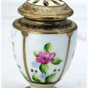 Może przedstawiać: Mały, zdobiony biało-złoty ceramiczny słoik z pokrywką. Słoik jest ozdobiony ręcznie malowanymi różowymi kwiatami i zielonymi liśćmi. Pokrywka ma uchwyt w kształcie pętli i dekoracyjne wycięcia. Podstawa i pokrywka są wykończone złotem.