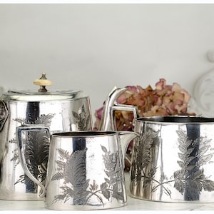 May include: A silver tea set comprising a teapot, creamer jug, and sugar bowl. Each item features a fern leaf pattern. The teapot has a light-coloured knob. The set is displayed on a white surface.