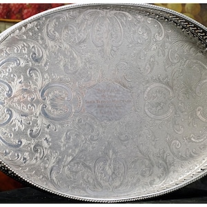May include: Oval silver serving tray with ornate, engraved floral patterns. The tray has a decorative, pierced border and a central engraved inscription. A classic piece for serving or display.