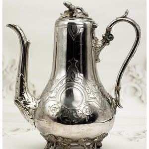 May include: A silver coffee pot with intricate detailing. The pot has a curved spout, a decorative handle, and a lid with a small finial. The body is engraved with floral and scroll patterns, and it stands on three decorative feet.