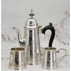 May include: A silver coffee set comprising a tall coffee pot with a black handle, a small creamer, and a tumbler. The set features engraved floral patterns and is displayed on a white surface.