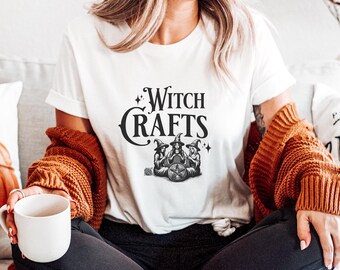 Könnte beinhalten: Weißes T-Shirt mit dem Text "Witch Crafts" in schwarzer, gotischer Schrift. Darunter eine Grafik von drei Hexen, die sich um ein Pentagramm versammeln. Die Hexen tragen spitze Hüte.