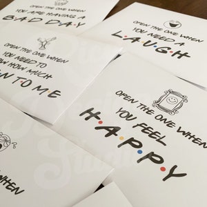 May include: Set of six white envelopes with black text and colorful dots. Each envelope has a different message: "Open the one when you are having a bad day", "Open the one when you need a laugh", "Open the one when you need to know how much I love you", "Open the one when you feel happy", "Open the one when you need a hug", and "Open the one when you need a smile".