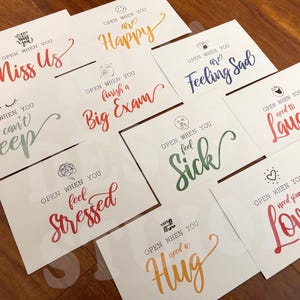May include: Ten colorful cards with different messages on them. Each card has the text "Open when you..." followed by a different feeling or situation. The cards are on a wooden surface.