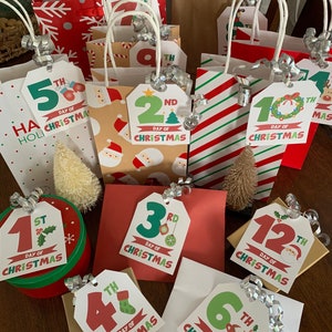 May include: A set of 12 Christmas gift bags with tags numbered 1st through 12th, each with a different Christmas-themed design. The bags are made of brown paper and have red and white snowflake patterns. The tags are decorated with holly, ornaments, and other festive elements.
