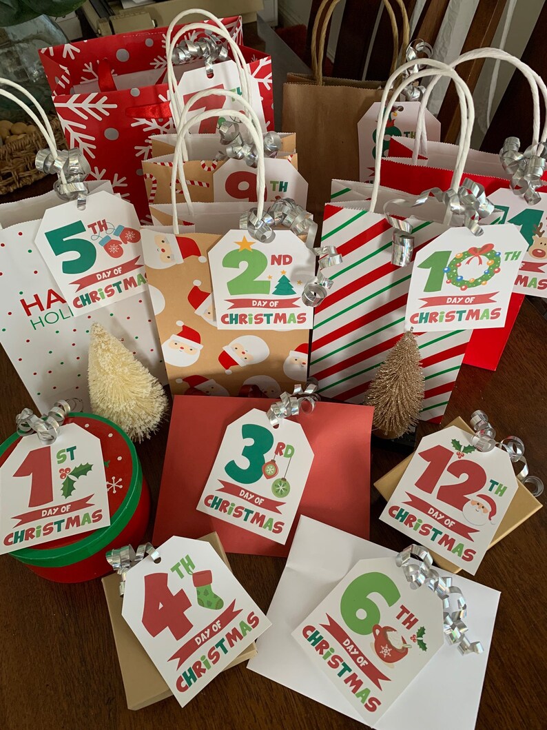 May include: A set of 12 Christmas gift bags with tags numbered 1st through 12th, each with a different Christmas-themed design. The bags are made of brown paper and have red and white snowflake patterns. The tags are decorated with holly, ornaments, and other festive elements.