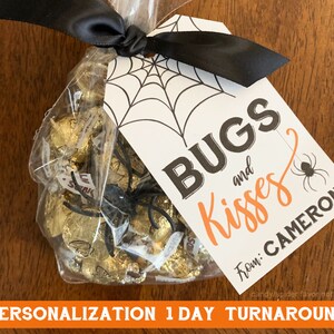 May include: A white tag with black spiderwebs and a black spider printed on it. The tag has the text "BUGS and Kisses" in black and orange lettering. Below the text is "From: CAMERON" in black lettering. The tag is tied to a clear plastic bag filled with gold-wrapped chocolate kisses.