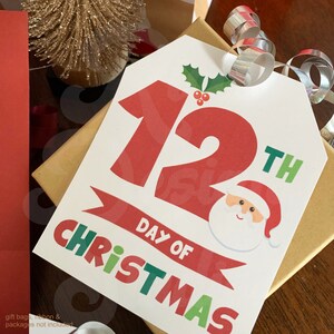 May include: White printable Christmas gift tag with red and green text. The tag says "12th Day of Christmas" with a cartoon Santa Claus image.