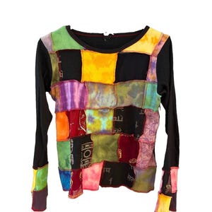 May include: A long-sleeve shirt featuring a patchwork design on the front panel. The garment is constructed with black fabric and various colorful, tie-dyed, and patterned fabric squares stitched together. It has a round neckline and long sleeves.