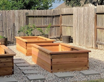 Op de afbeelding: Drie verhoogde tuinbedden gemaakt van cederhout. De bedden bevinden zich in een tuin met een houten hek op de achtergrond.