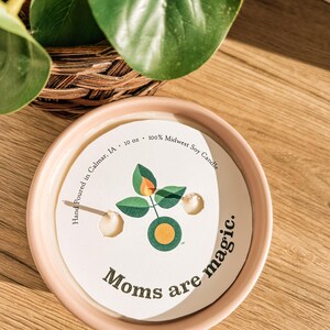 May include: A round, light pink candle holder with a white label that reads "Moms are magic." The label features a green and orange graphic. The candle is 10 oz and made with 100% Midwest Soy.