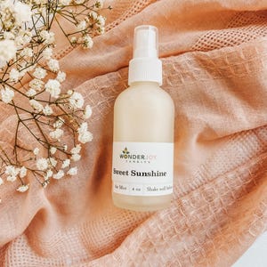 May include: A white spray bottle with a clear cap, labeled "Sweet Sunshine" air mist, sits on a peach-colored textured fabric. Delicate white flowers are arranged on the left side of the image, adding a soft, natural touch.