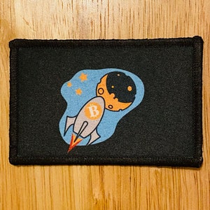 May include: A black patch with a white rocket ship flying towards a crescent moon with a 'B' on the side of the rocket. The moon is orange and the background is blue with white stars.