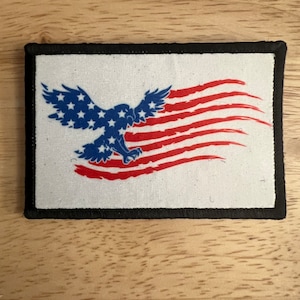May include: A white patch with a black border featuring a red, white, and blue American flag with an eagle flying through it.