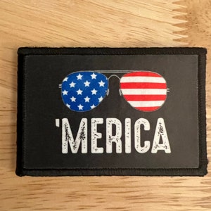May include: A black patch with a white border featuring a pair of sunglasses with an American flag design and the text 'MERICA' in white with a distressed look.