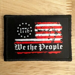 May include: A black patch with a distressed American flag design. The flag is red, white, and blue with the words "We the People" in white text below the flag. The year "1776" is in white text inside a circle of stars above the flag.