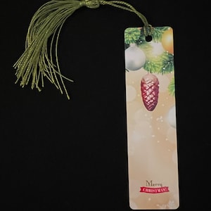 May include: A rectangular Christmas-themed bookmark with a green tassel. The bookmark features festive illustrations of ornaments, pine needles, and a pinecone. The words "Merry Christmas!" are printed on the bottom.