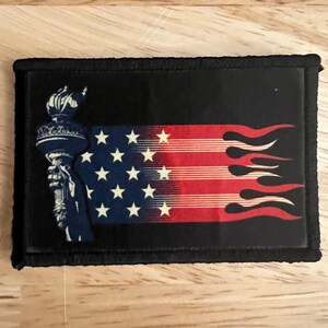 May include: A black patch with a white and red American flag with flames on the right side and the Statue of Liberty on the left side.