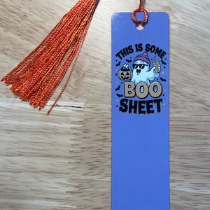 May include: A blue bookmark with an orange tassel. The bookmark features a cartoon ghost wearing sunglasses and a beanie, holding a pumpkin and a drink. Text on the bookmark reads "THIS IS SOME BOO SHEET."