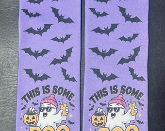 May include: A pair of purple Halloween socks with a pattern of black bats. The socks feature a cartoon ghost wearing sunglasses and a pink beanie, holding a pumpkin and a drink. The text "THIS IS SOME BOO SHEET" is printed on the socks.