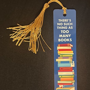 May include: Blue bookmark with the text "THERE'S NO SUCH THING AS TOO MANY BOOKS" and a stack of colorful books. A gold tassel hangs from the top. The bookmark is rectangular and has a glossy finish.