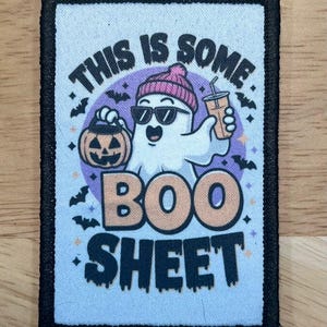 May include: A rectangular patch with a black border features a cartoon ghost wearing sunglasses and a pink beanie, holding a pumpkin and a drink. The text "THIS IS SOME BOO SHEET" is displayed in various colors and fonts, with bats and stars.