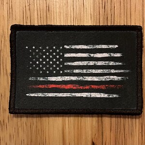 May include: A black and white American flag patch with a red stripe across the middle. The flag is distressed and has a worn look.