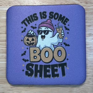 May include: A square purple coaster with a Halloween-themed design. The design features a cartoon ghost wearing sunglasses and a beanie, holding a pumpkin and a drink. The text reads "THIS IS SOME BOO SHEET."