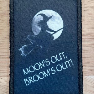 May include: Black patch with a witch silhouetted against a full moon. The text "MOON'S OUT, BROOM'S OUT!" is printed in white. The patch has a black border.