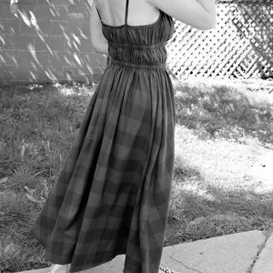 May include: A woman wearing a black and white plaid maxi dress with a gathered waist and thin straps. She is wearing black sandals with silver studs.