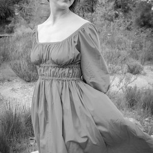 May include: A woman in a long, flowing, pink dress stands in a field of tall grass. The dress has a gathered waist and a square neckline. The woman is looking off to the side, and her hair is blowing in the wind.