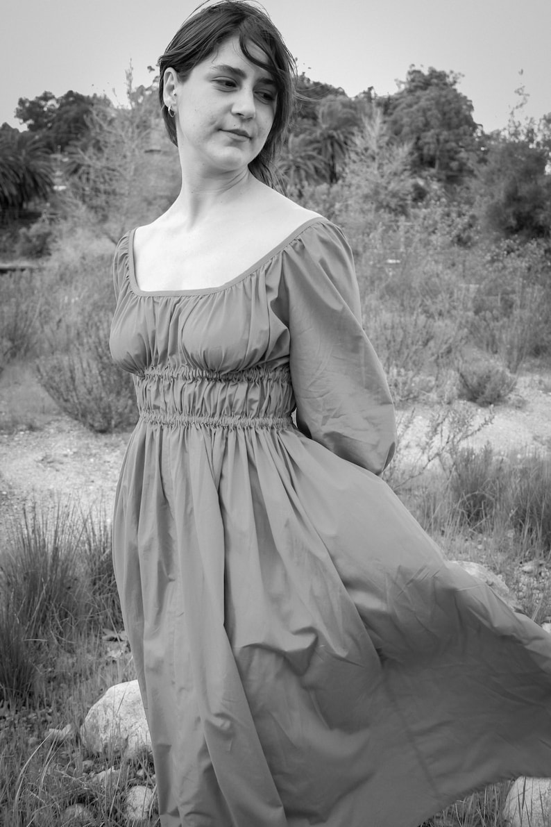 May include: A woman in a long, flowing, pink dress stands in a field of tall grass. The dress has a gathered waist and a square neckline. The woman is looking off to the side, and her hair is blowing in the wind.