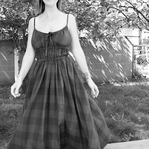 May include: A woman in a black and white plaid dress smiles while standing on a sidewalk. The dress has a fitted bodice and a flowing skirt.
