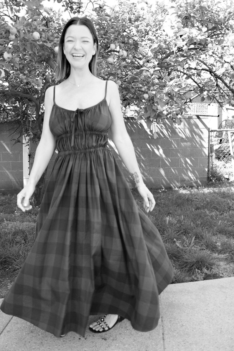 May include: A woman in a black and white plaid dress smiles while standing on a sidewalk. The dress has a fitted bodice and a flowing skirt.