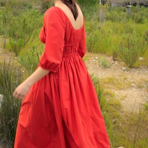May include: A woman wearing a red, long-sleeved dress with a gathered waist and a square neckline. The dress is flowing in the wind as she walks through a grassy field.