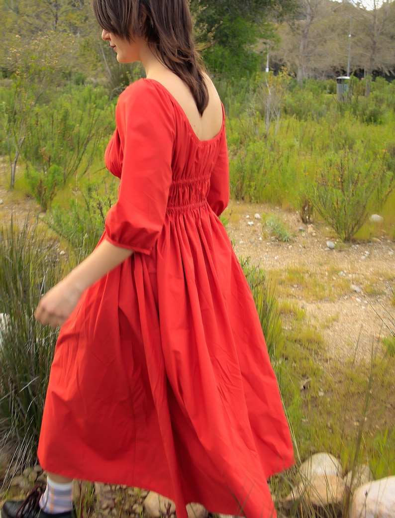 May include: A woman wearing a red, long-sleeved dress with a gathered waist and a square neckline. The dress is flowing in the wind as she walks through a grassy field.