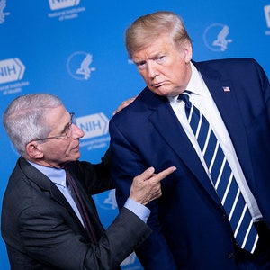 May include: A man in a blue suit and a striped tie stands with his arm around another man in a gray suit and a purple tie. The man in the blue suit is looking to the right, while the man in the gray suit is looking at him and pointing to his chest. The background is a blue wall with the NIH logo.