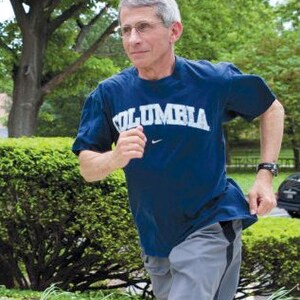 May include: A man wearing a blue t-shirt with the word "COLUMBIA" printed on it in white letters. He is running outdoors, wearing grey shorts and a black watch.