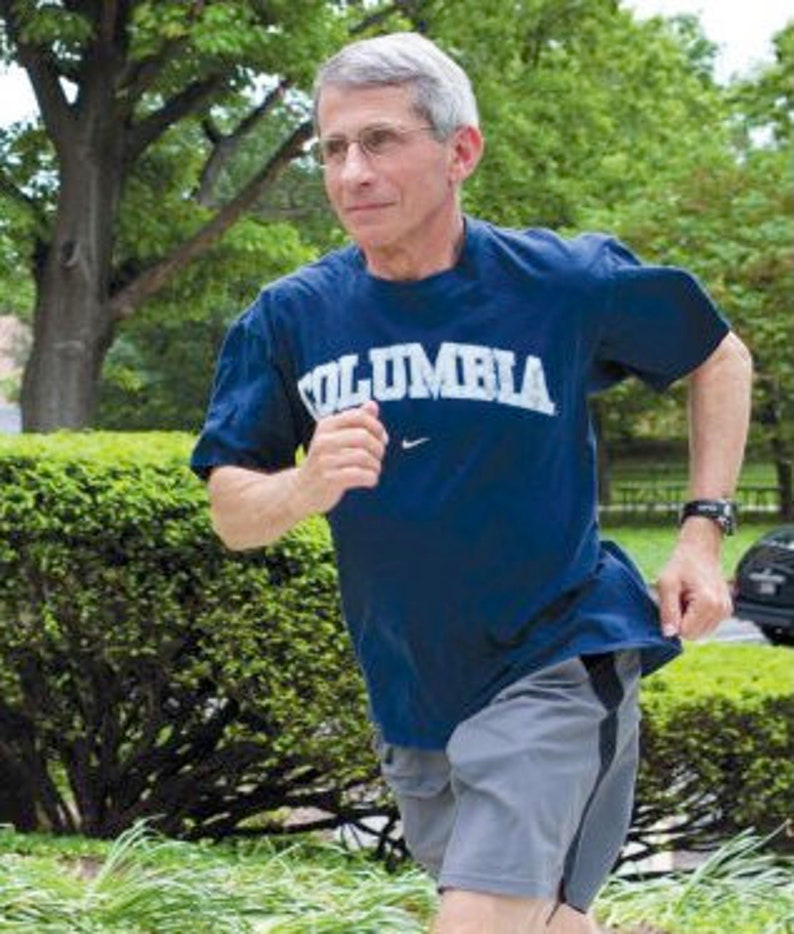 May include: A man wearing a blue t-shirt with the word "COLUMBIA" printed on it in white letters. He is running outdoors, wearing grey shorts and a black watch.
