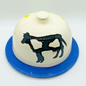 May include: A ceramic butter dish with a blue base and a domed lid. The lid is off-white with a black cow silhouette and yellow star accents. The dish is designed for serving butter.