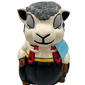 May include: A ceramic sheep figurine with a cream-colored face, gray wool, and black ears. It wears black overalls with red suspenders and a yellow waistband. The sheep has closed eyes and red flower accents.