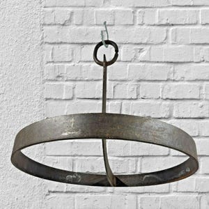 May include: A rustic, circular metal hanging pot rack with a central support and a ring for hanging. The rack is made of weathered, dark metal and is suspended from a chain against a white brick wall.